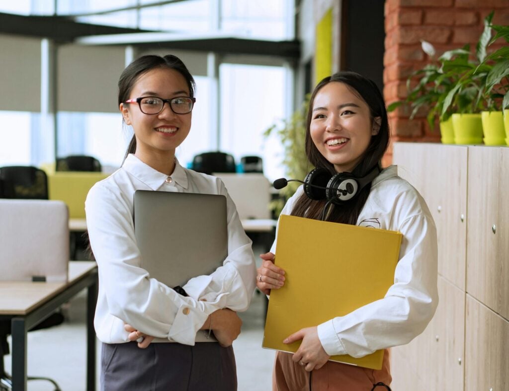 Two smiling Asian women in a modern office, holding documents and laptop, showcasing teamwork.